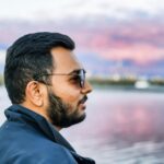 Bearded man in sunglasses gazing at sunset over tranquil lake, showcasing serene and thoughtful atmosphere.